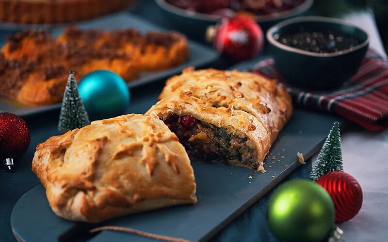 Auf einem weihnachtlich gedeckten Tisch mit bunten Kugeln steht eine blaue Platte mit einem veganen Pilz-Rindfleisch Wellington. Es ist in der Mitte durchgeschnitten. Im Hintergrund sind verschwommen weitere Speisen zu sehen.