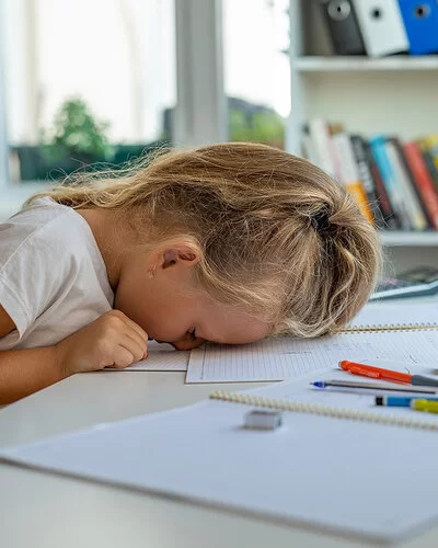 Ein kleines Mädchen mit langen blonden Haaren sitzt an einem weißen Tisch vor ihren Schulaufgaben und hat den Kopf mit der Stirn auf ihr Heft gelegt.