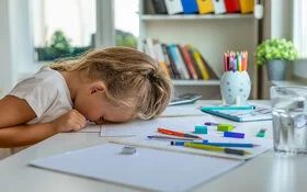 Ein kleines Mädchen mit langen blonden Haaren sitzt an einem weißen Tisch vor ihren Schulaufgaben und hat den Kopf mit der Stirn auf ihr Heft gelegt.