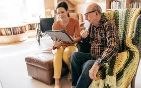 Eine weibliche Pflegekraft sitzt zusammen mit einem alten Mann in dessen Wohnzimmer, im Hintergrund steht ein Rollstuhl. Die Pflegerin hält ein Tablet in den Händen und beide schauen auf den Bildschirm.