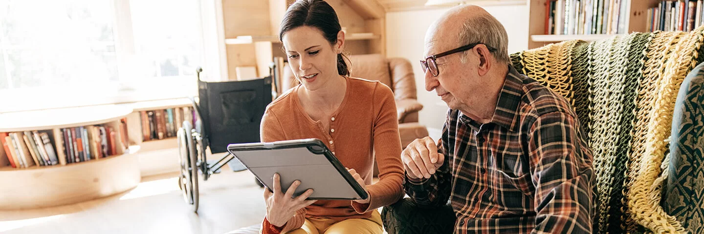 Eine weibliche Pflegekraft sitzt zusammen mit einem alten Mann in dessen Wohnzimmer, im Hintergrund steht ein Rollstuhl. Die Pflegerin hält ein Tablet in den Händen und beide schauen auf den Bildschirm.