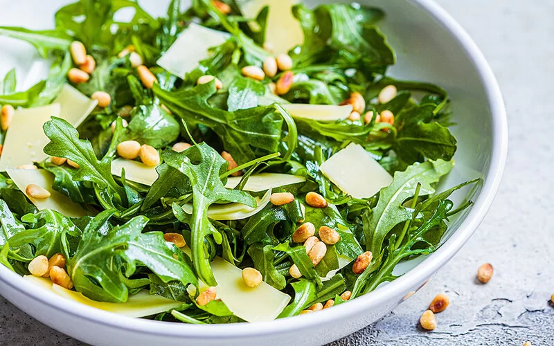 Nahaufnahme einer weißen Schüssel mit Rucola-Blättern, Pinienkernen und gehobeltem Parmesan.