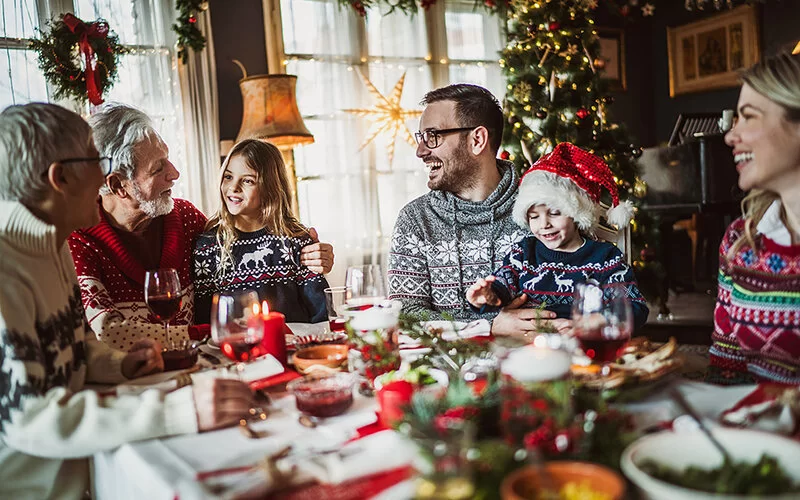 Drei Generationen sitzen an einem Esstisch in einem weihnachtlich dekorierten Raum und lachen gemeinsam.