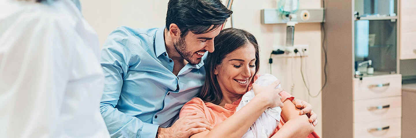 Eine junge Familie auf der Geburtsstation im Krankenhaus. Die Frau hält ihr neugeborenes Baby im Arm.