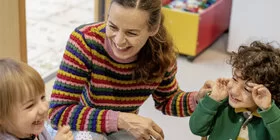 Zwei Kinder sitzen mit einer Erzieherin lachend an einem Tisch und essen ihr Brot. Das Kita-Programm JolinchenKids der AOK fördert die Gesundheit von Kindern.