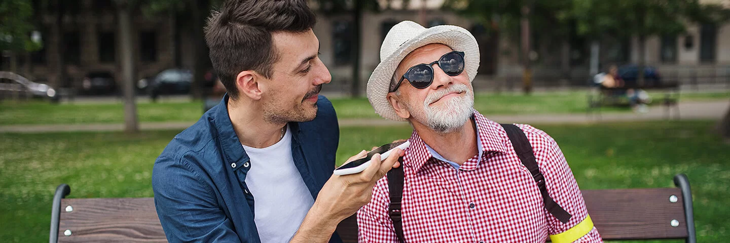 Ein junger Mann und ein älterer mit Sehbehinderung sitzen im Park auf einer Bank. Der junge Mann richtet ein Smartphone zum rechten Ohr des älteren Mannes. Dieser schmunzelt über das, was er hört.