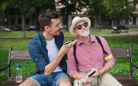 Ein junger Mann und ein älterer mit Sehbehinderung sitzen im Park auf einer Bank. Der junge Mann richtet ein Smartphone zum rechten Ohr des älteren Mannes. Dieser schmunzelt über das, was er hört.