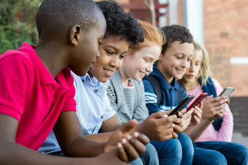 Fünf Jungen und Mädchen aus der dritten oder vierten Klassenstufe sitzen nebeneinander und schauen auf Smartphones.