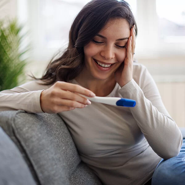 Eine junge Frau mit langen dunklen Haaren sitzt auf einem grauen Sofa und schaut lächelnd auf einen Schwangerschaftstest, den sie in der Hand hält.