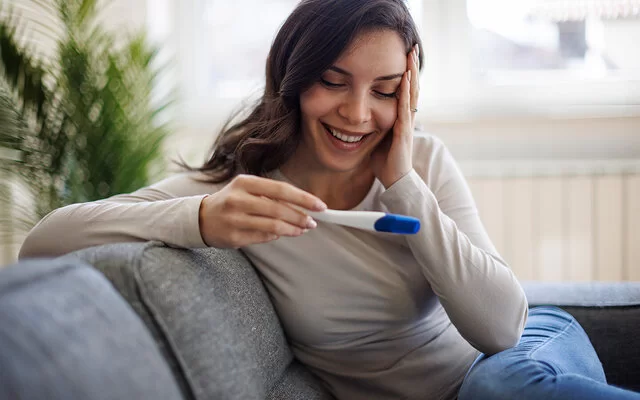 Eine junge Frau mit langen dunklen Haaren sitzt auf einem grauen Sofa und schaut lächelnd auf einen Schwangerschaftstest, den sie in der Hand hält.