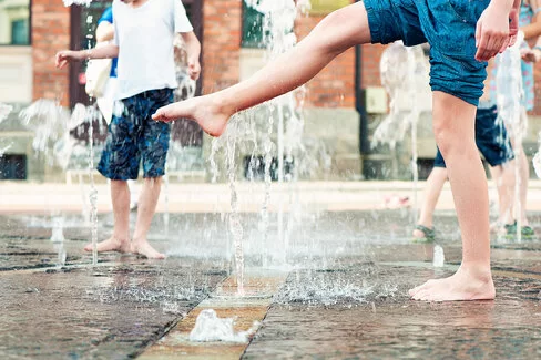 Mehrere Kinder laufen barfuß in kurzen Hosen in einem öffentlichen Springbrunnen herum.