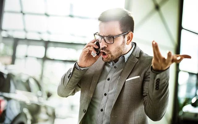 Ein Geschäftsmann in Hemd und Sakko telefoniert in einem Büro. Er schreit wütend in sein Smartphone und gestikuliert mit der linken Hand.