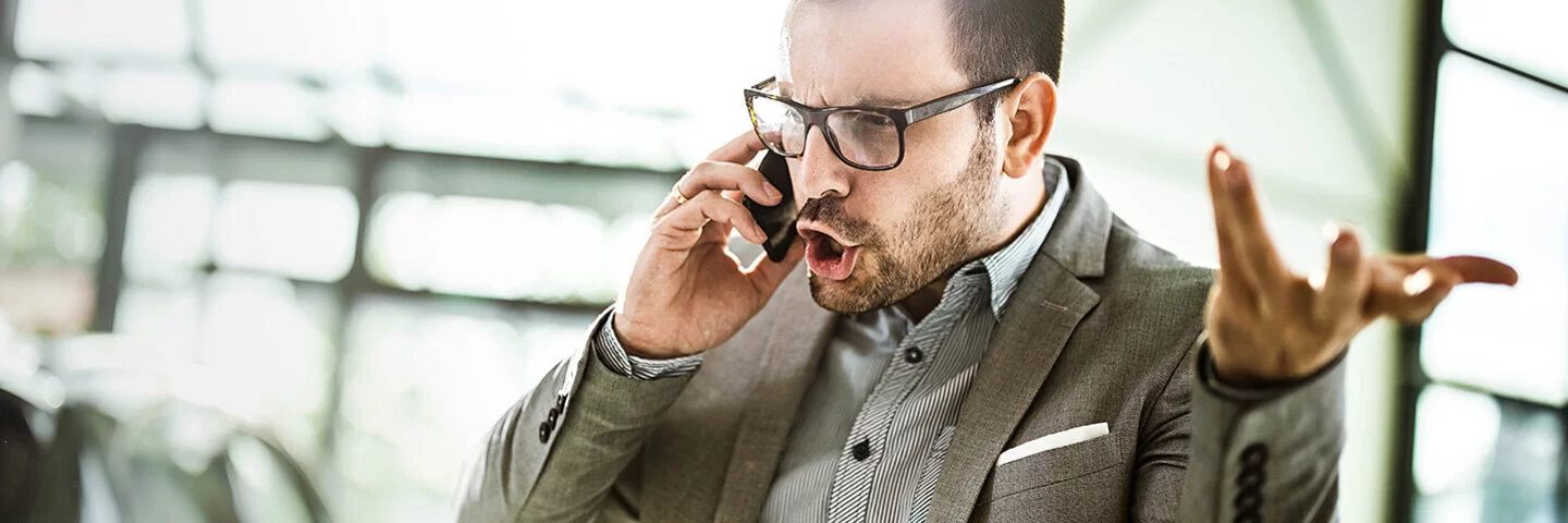 Ein Geschäftsmann in Hemd und Sakko telefoniert in einem Büro. Er schreit wütend in sein Smartphone und gestikuliert mit der linken Hand.