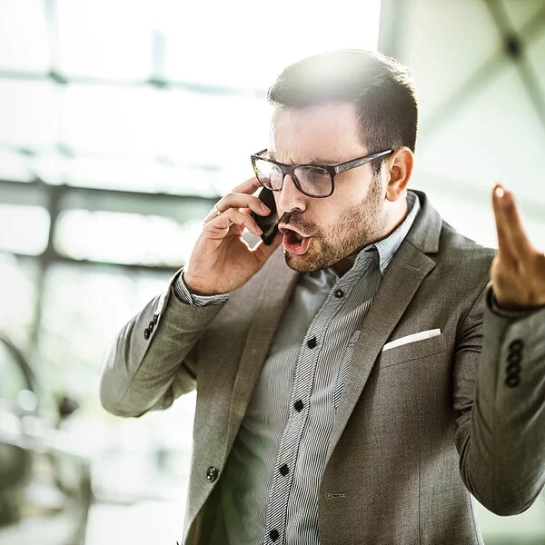 Ein Geschäftsmann in Hemd und Sakko telefoniert in einem Büro. Er schreit wütend in sein Smartphone und gestikuliert mit der linken Hand.