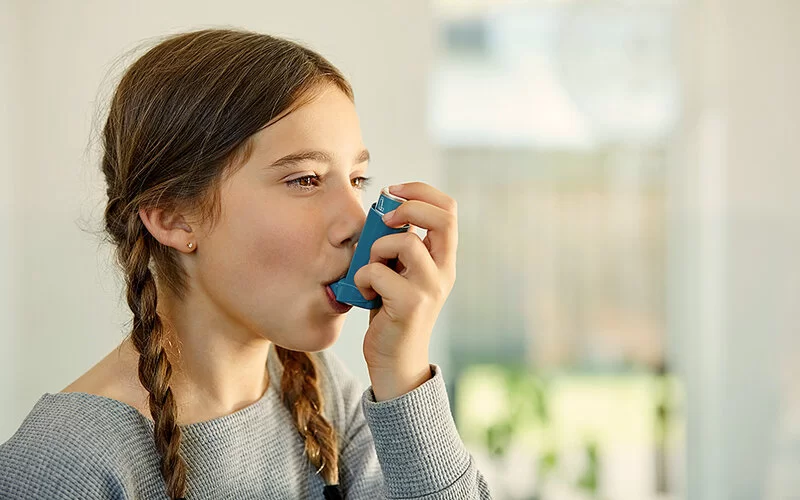 Ein etwa 9-jähriges Mädchen mit geflochtenen Zöpfen ist von der Seite zu sehen. Sie ist zuhause und benutzt einen Asthma-Inhalator.