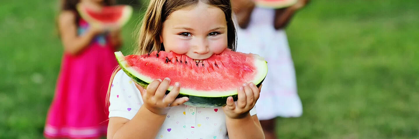 Ein kleines Mädchen steht in einem weißen Sommerkleid mit bunten Herzchen auf einer Wiese in einem Park und beißt genießerisch in eine Wassermelonenspalte. Hinter ihr sind zwei weitere Mädchen in bunten Kleidern zu sehen.