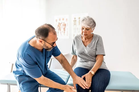 Eine ältere Frau sitzt in einem Behandlungsraum auf einer Behandlungsliege. Ein Orthopäde tastet ihr rechtes Knie ab.