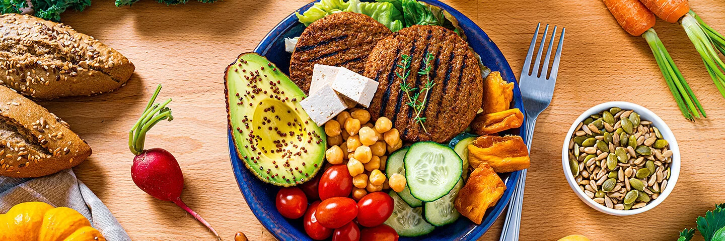 Auf einem Tisch steht ein Teller mit gesundem und veganem Essen: Tomaten, Kichererbsen, Avocado, Gurken, Salat, getrocknetes Gemüse und pflanzliche Burger Pattys. Daneben liegen Möhren, Bohnen, rote Paprika, Kartoffeln, Kürbis, ein Radieschen, Tomaten und Petersilie. In einer kleinen Schüssel sind Kürbiskerne enthalten.