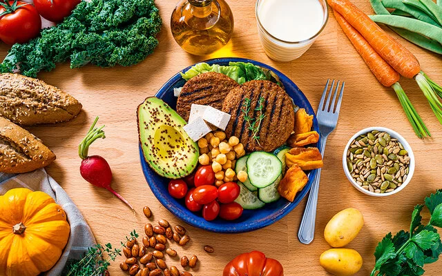 Auf einem Tisch steht ein Teller mit gesundem und veganem Essen: Tomaten, Kichererbsen, Avocado, Gurken, Salat, getrocknetes Gemüse und pflanzliche Burger Pattys. Daneben liegen Möhren, Bohnen, rote Paprika, Kartoffeln, Kürbis, ein Radieschen, Tomaten und Petersilie. In einer kleinen Schüssel sind Kürbiskerne enthalten.