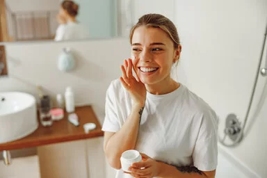 Eine junge Frau steht im Badezimmer und cremt sich lächelnd ihr Gesicht ein.