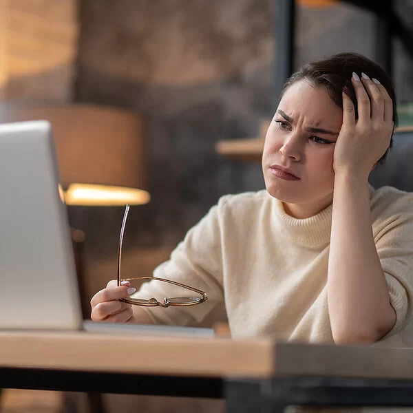 Eine Frau sitzt an einem Tisch und schaut sorgenvoll auf den Bildschirm eines Laptops. In der einen Hand hält sie eine Brille.