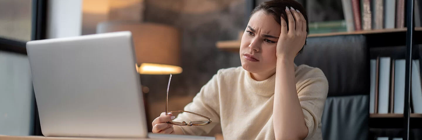 Eine Frau sitzt an einem Tisch und schaut sorgenvoll auf den Bildschirm eines Laptops. In der einen Hand hält sie eine Brille.