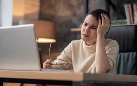 Eine Frau sitzt an einem Tisch und schaut sorgenvoll auf den Bildschirm eines Laptops. In der einen Hand hält sie eine Brille.