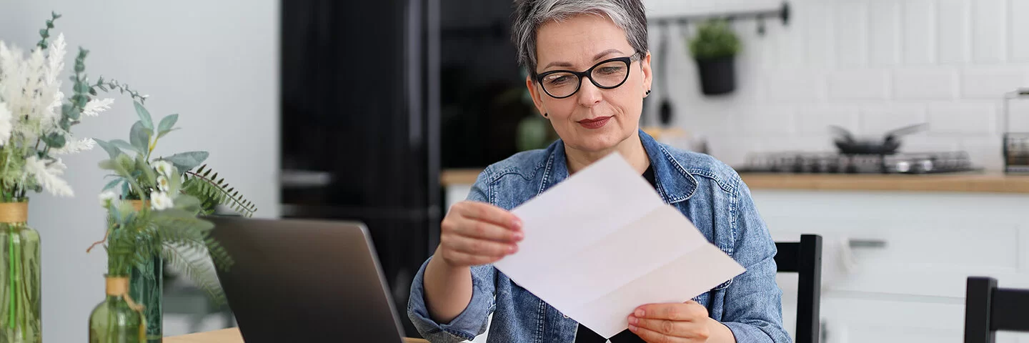 Eine Frau mittleren Alters mit kurzen Haaren, einige davon bereits grau, sitzt in einer Küche an einem Esstisch. Sie liest einen Brief, den sie in ihren Händen hält. Auf dem Tisch vor ihr steht ein Laptop.