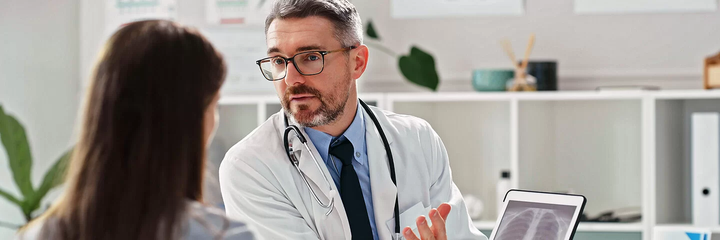 Das Bild zeigt einen Arzt mittleren Alters mit grauen Strähnen in Vollbart und Haaren,  der einer Patientin gegenüber sitzt und ihr ihre Röntgenaufnahmen auf einem digitalen Tablet zeigt.