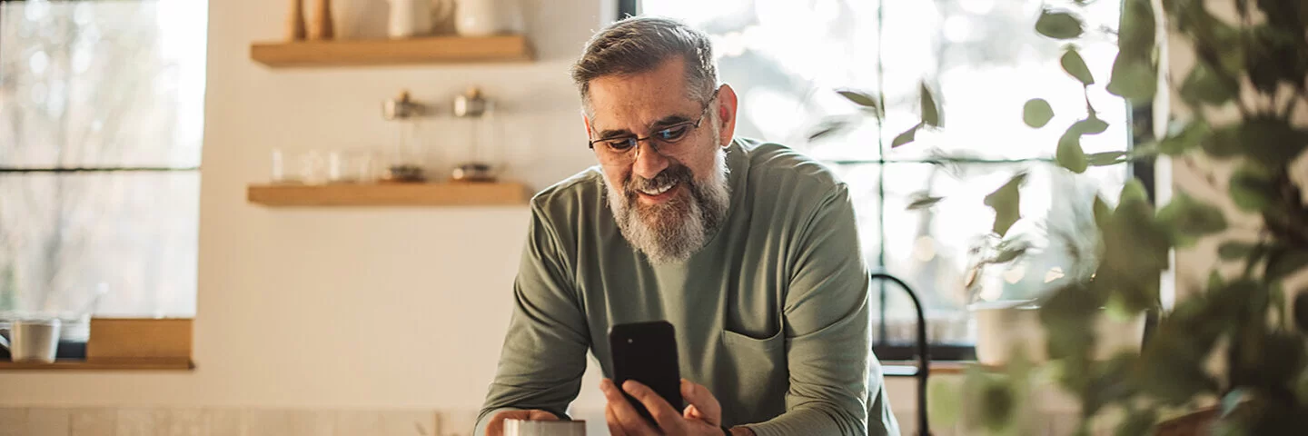 Ein Mann mittleren Alters steht in einer Küche und blickt auf ein Smartphone in seiner linken Hand.