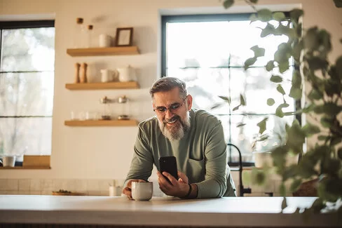 Ein Mann mittleren Alters steht in einer Küche und blickt auf ein Smartphone in seiner linken Hand.