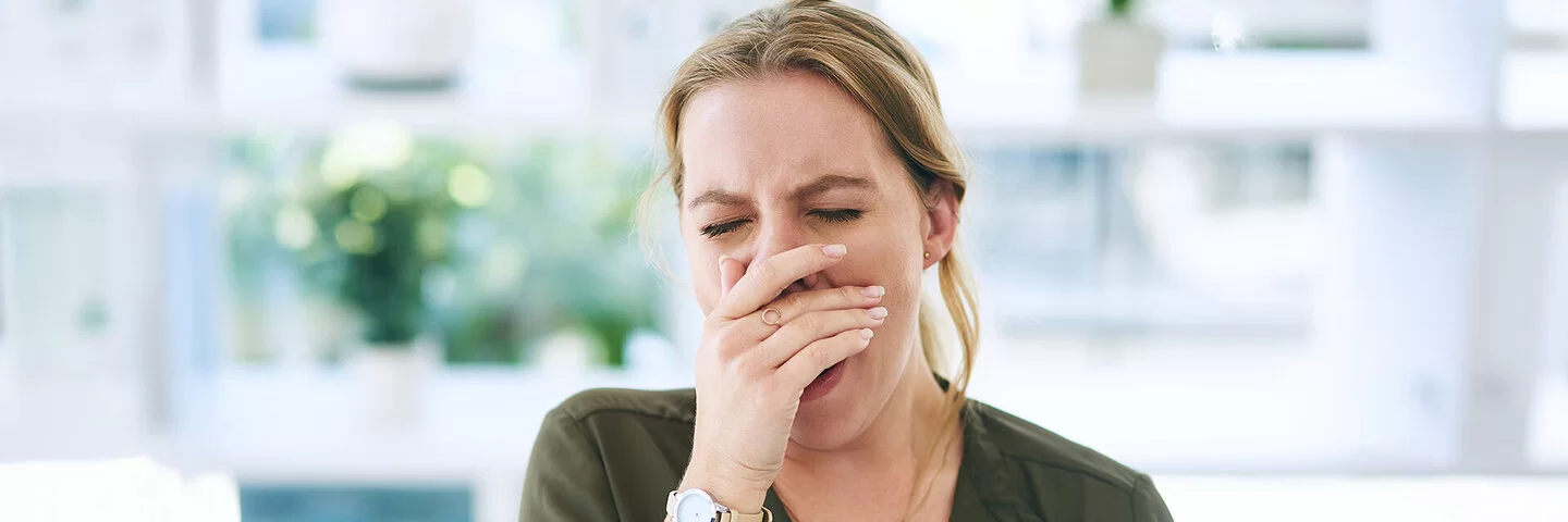 Eine junge Frau in einem lichtdurchfluteten Raum gähnt. Sie hält sich die Hand vor den Mund.