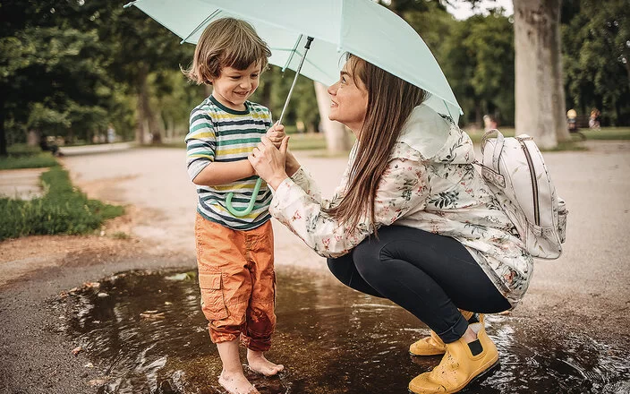 Eine junge Mutter mit ihrem Sohn lächelnd unter einem Regenschirm in einer Pfütze.