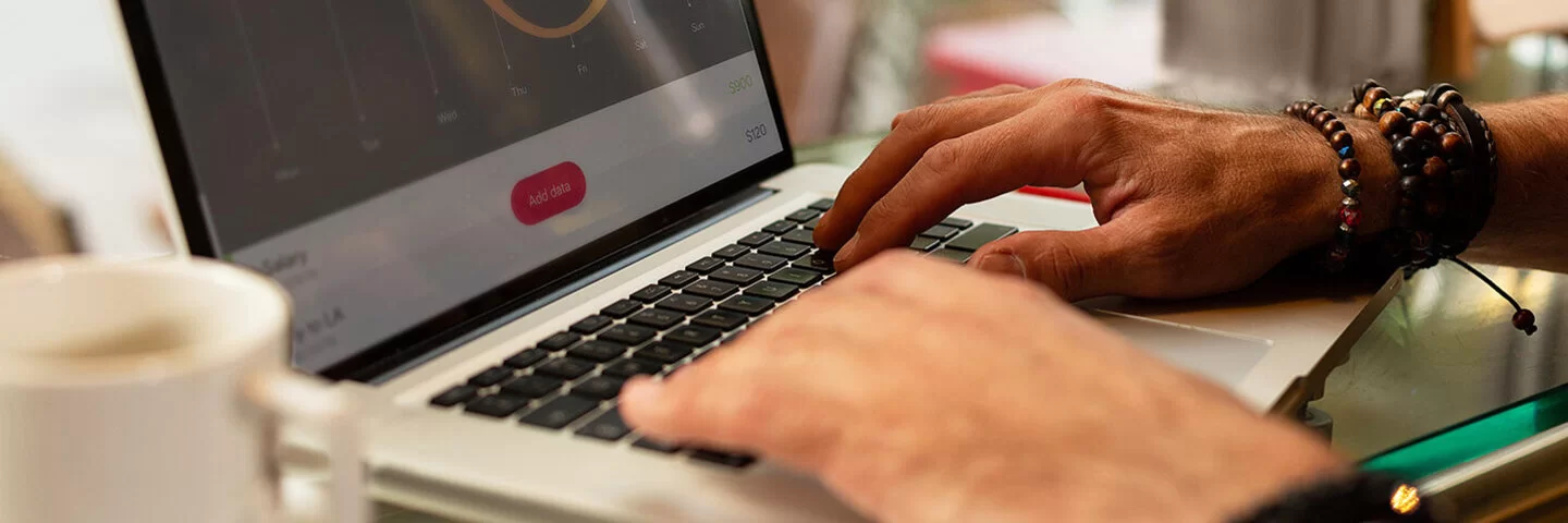 Der Bildschirm eines Laptops mit einem Aktienkurs-Diagramm und zwei Hände einer Person. Eine Hand liegt auf der Tastatur, die andere zeigt auf eine Zahl auf dem Bildschirm.