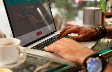 Der Bildschirm eines Laptops mit einem Aktienkurs-Diagramm und zwei Hände einer Person. Eine Hand liegt auf der Tastatur, die andere zeigt auf eine Zahl auf dem Bildschirm. 