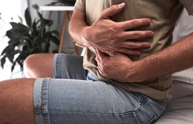 Ein sitzender Mann in einem olivgrünen T-Shirt hält sich mit den Händen die linken Rippen.