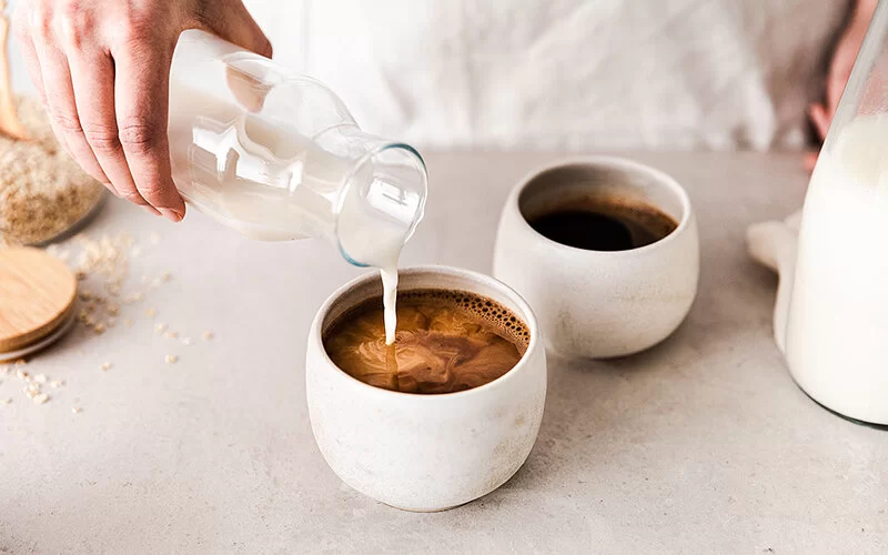 Eine nicht sichtbare Person gießt Haferdrink aus einer Glaskaraffe in eine Tasse mit Kaffee.