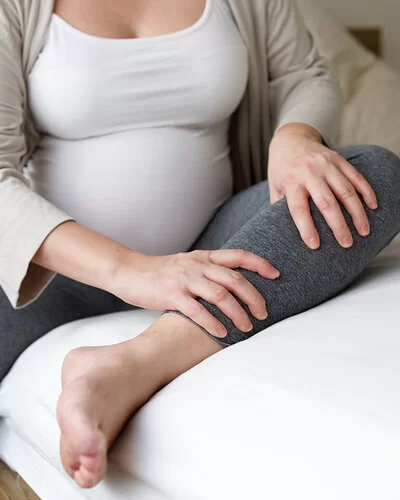 Eine schwangere Frau sitzt auf einem Bett. Ihr Gesicht ist nicht zu sehen. Das rechte Bein ist auf dem Boden aufgestellt, das linke liegt nach innen angewinkelt auf dem Bett. Mit den Händen massiert die Schwangere ihren linken Unterschenkel.