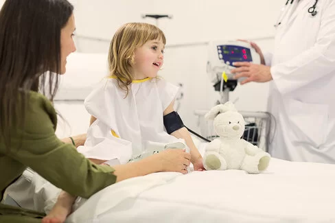 Ein junges Mädchen in weißer Patientenkleidung sitzt aufrecht auf einem Krankenhausbett, während ihr Blutdruck gemessen wird. Neben ihr sitzt ihre Mutter in einem grünen Pullover.