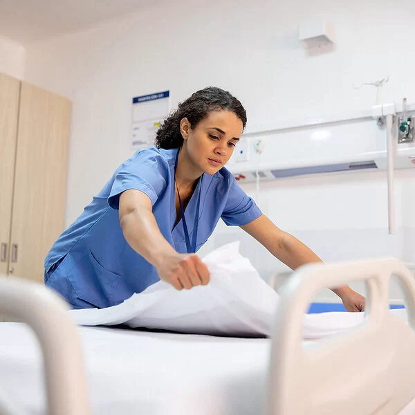 Eine junge Pflegefachfrau in blauem Kittel bereitet in einem Krankenhaus ein Patientenbett auf.