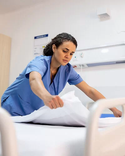 Eine junge Pflegefachfrau in blauem Kittel bereitet in einem Krankenhaus ein Patientenbett auf.