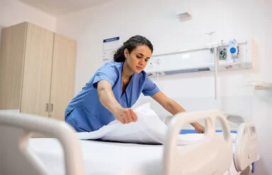 Eine junge Pflegefachfrau in blauem Kittel bereitet in einem Krankenhaus ein Patientenbett auf.