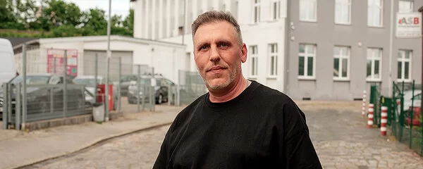 Ein mittelalter Mann in schwarzem T-Shirt und mit Tattoo am rechten Arm steht auf einer Straße und blickt in die Kamera.