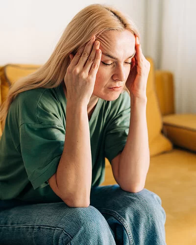 Eine Frau sitzt auf einem gelben Sofa und fasst sich an den Kopf. Sie hat starke Kopfschmerzen.