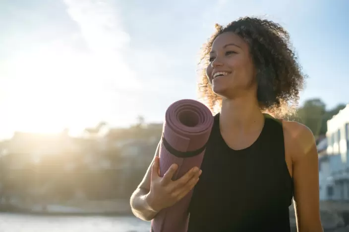 Eine junge Frau lächelt in der Sonne und hält eine Yogamatte unter dem Arm.