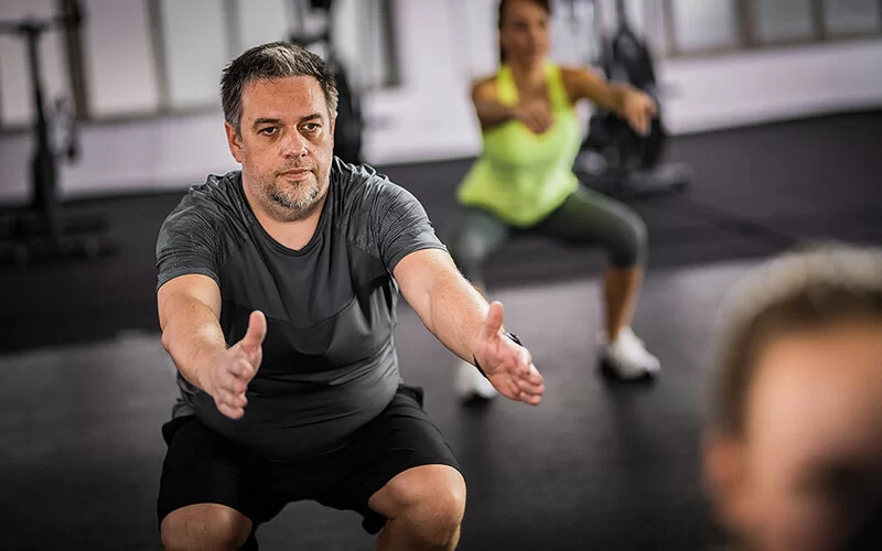 Ein leicht übergewichtiger Mann mit angestrengtem Gesichtsausdruck macht Kniebeugen im Fitnessstudio.