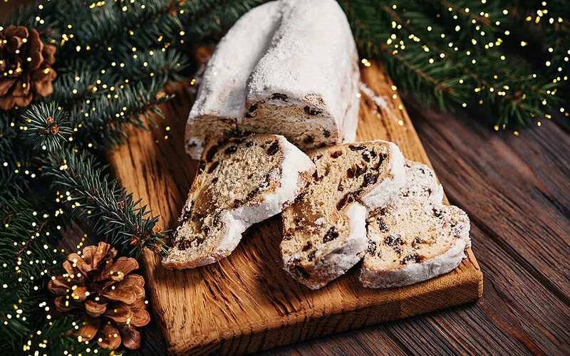 Ein aufgeschnittener Weihnachtsstollen mit drei Scheiben liegt auf einem Holzbrett. Der Stollen enthält Rosinen und ist von außen mit Puderzucker bedeckt. Die Umgebung des Holzbrettes ist weihachtlich mit Tannenzweigen, Tannenzapfen und Lichterketten geschmückt.