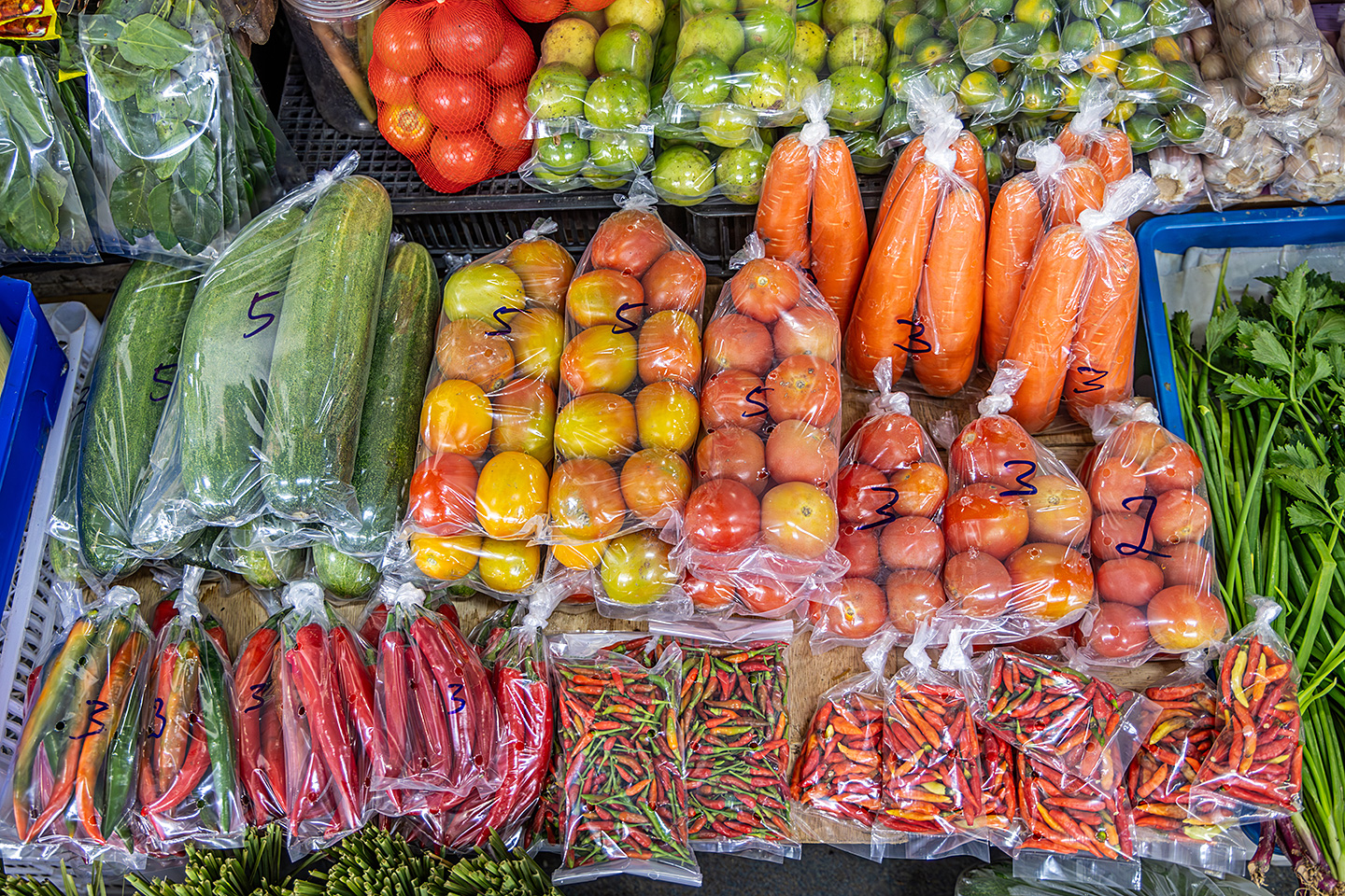 Nahaufnahme der Auslage eines Gemüsemarktstandes mit unter anderem Gurken, Tomaten, Karotten und Peperoni. Das Gemüse wird in Kunststoffverpackungen angeboten.