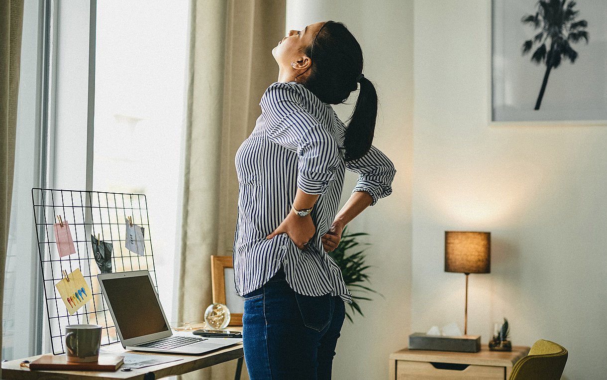 Foto einer jungen Frau, die im Homeoffice vor ihrem Laptop steht und sich schmerzverzerrt an den Rücken fasst.