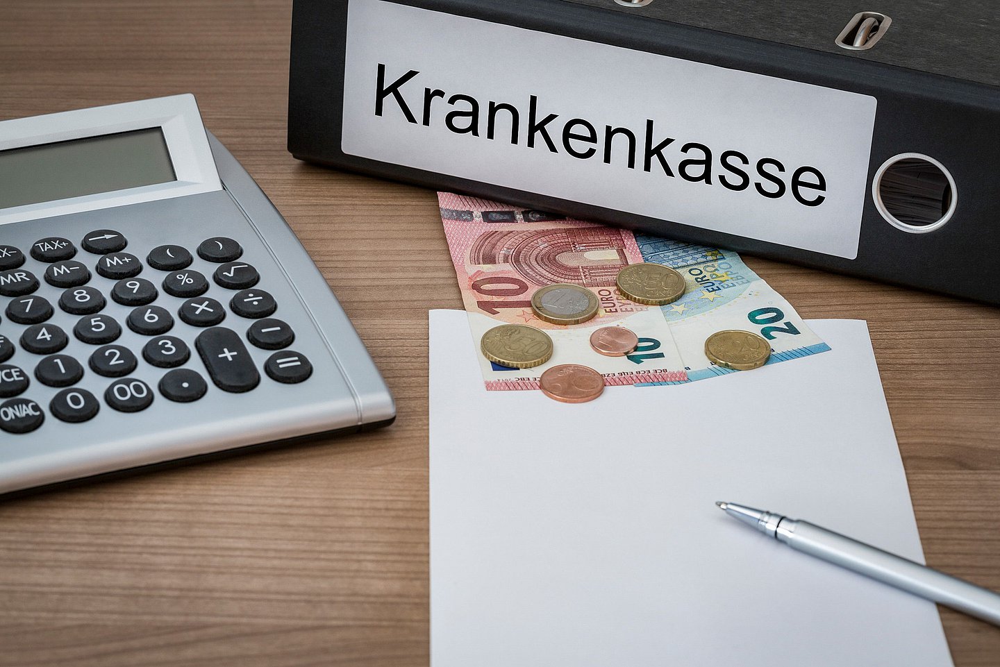 Auf einem Schreibtisch liegt ein Ordner mit der Aufschrift 'Krankenkasse', ein Taschenrechner und Geld.